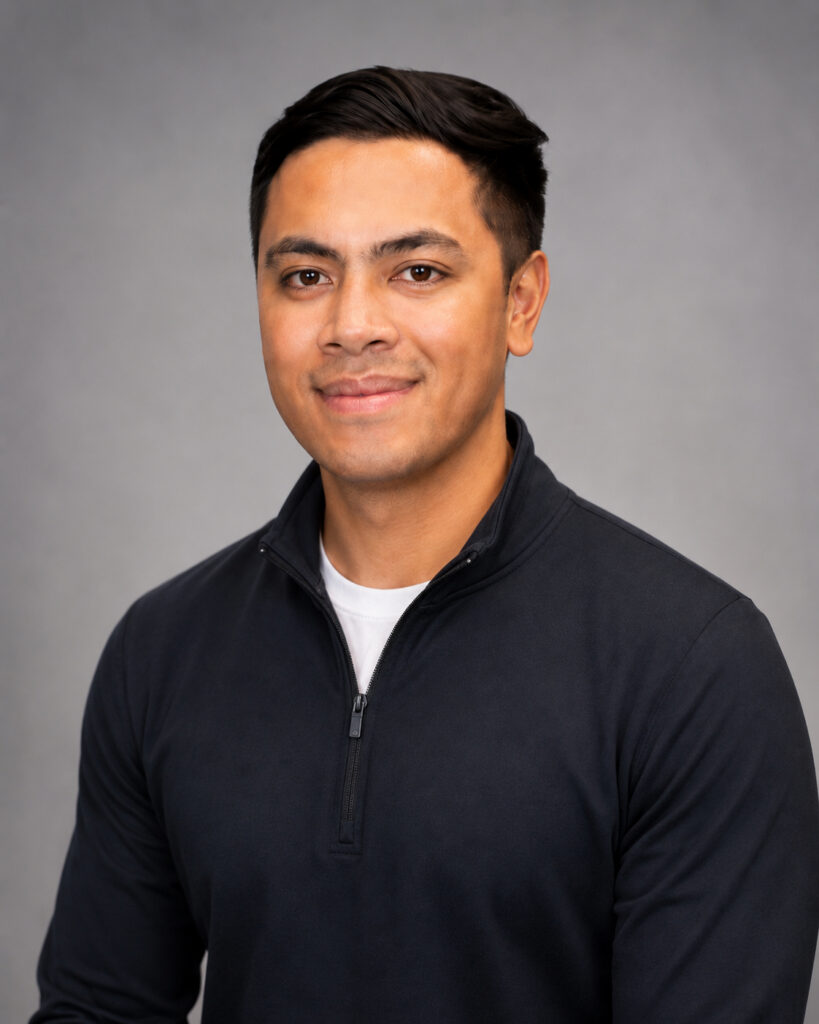 Professional headshot of a smiling man with short black hair, wearing a dark zip-neck sweater over a white tee against a gray studio background.